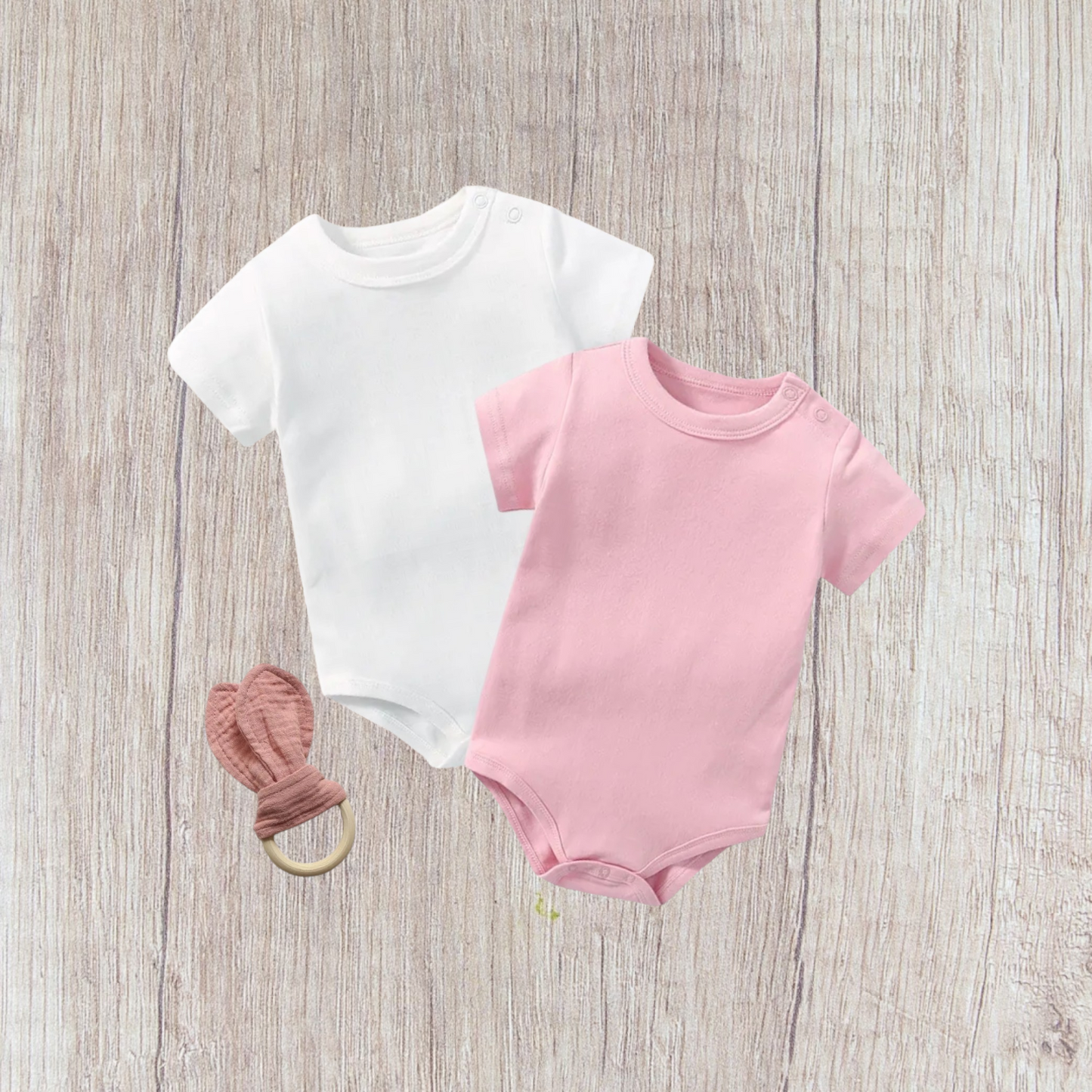 Baby bodysuit set with white and pink bodysuits and rabbit ear teether on wooden background.