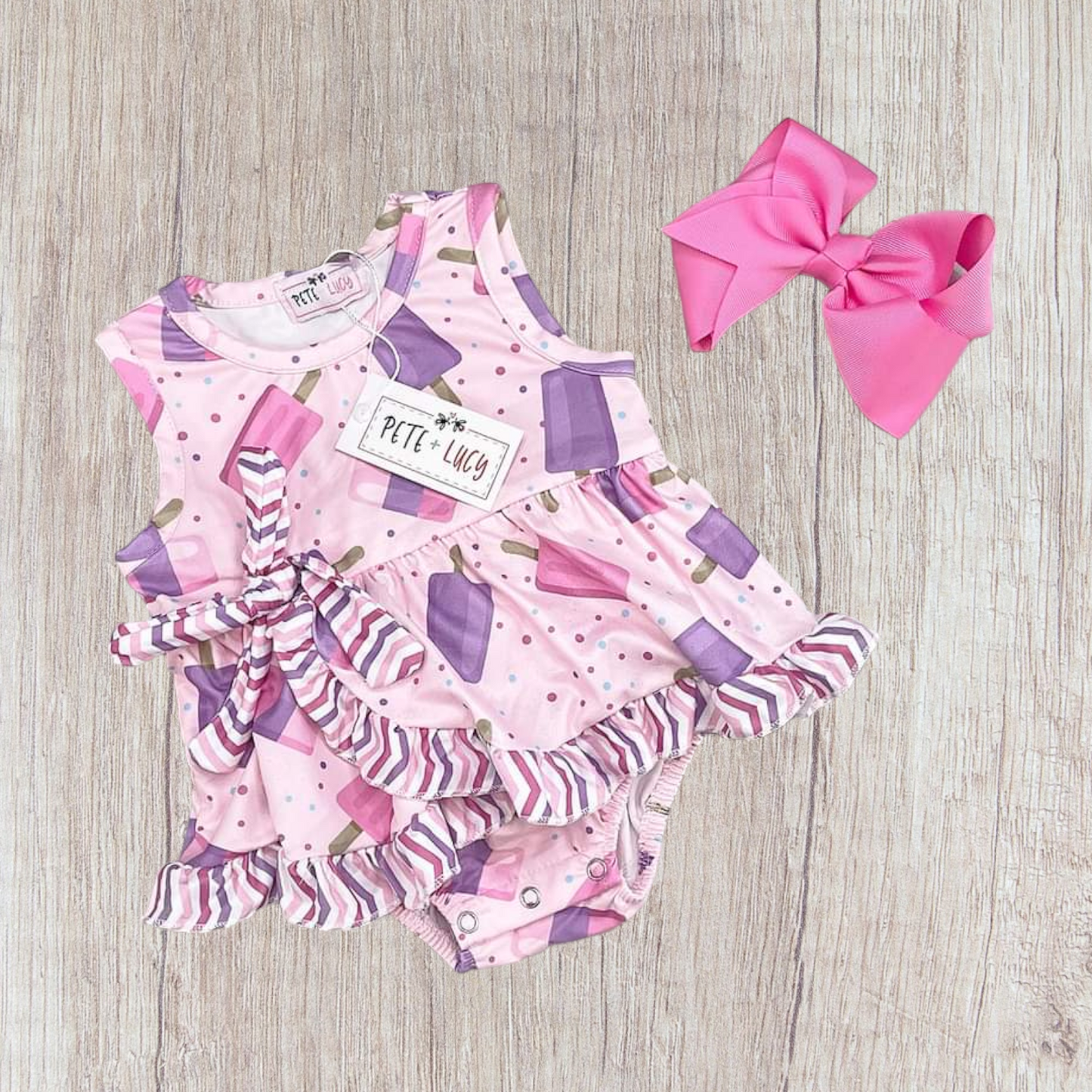 Popsicle Princess Romper with pink and purple popsicle print, ruffle details, and a side bow, shown on a light wood background.