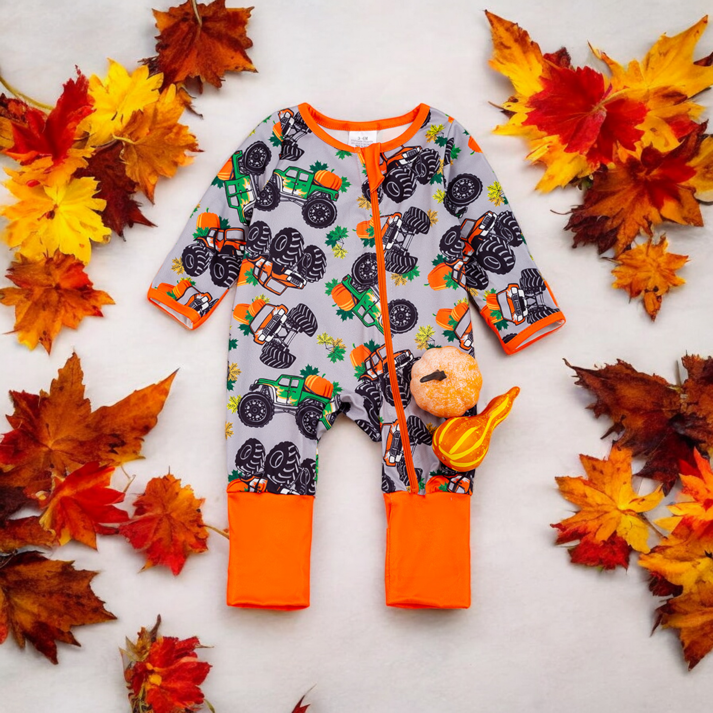 Baby romper with fall monster truck, pumpkins, and leaves pattern, cozy orange cuffs, and front zipper surrounded by autumn leaves and pumpkins.