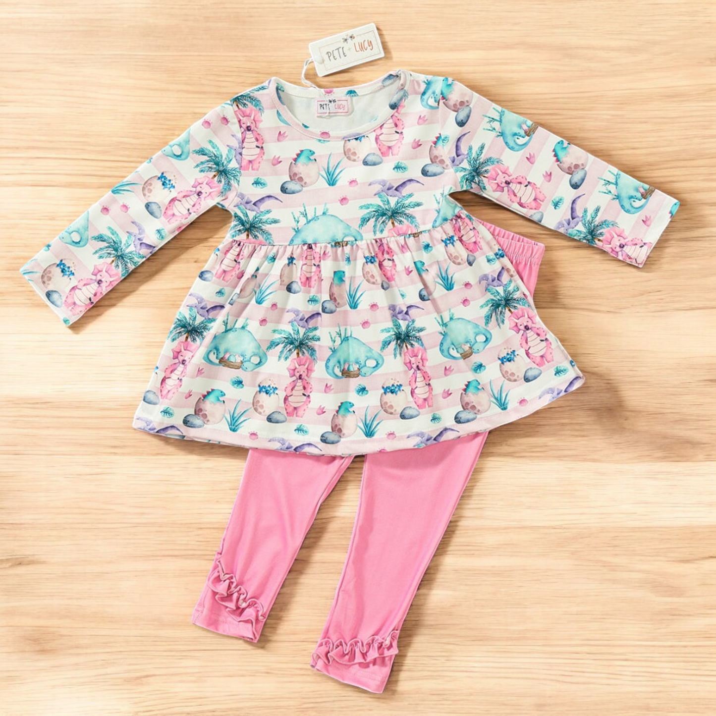 Mia Dinosaur 2 Piece Pant Set with tunic featuring dinosaur and palm tree print and pink ruffle leggings on a wooden background.