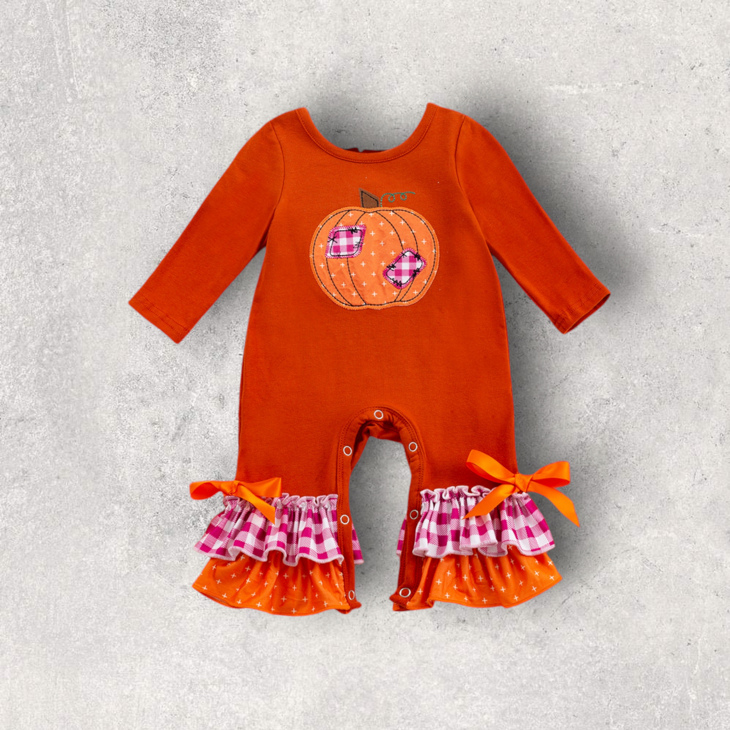Orange baby romper with pumpkin appliqué, plaid and polka-dot ruffle legs, and orange bows, perfect for fall.