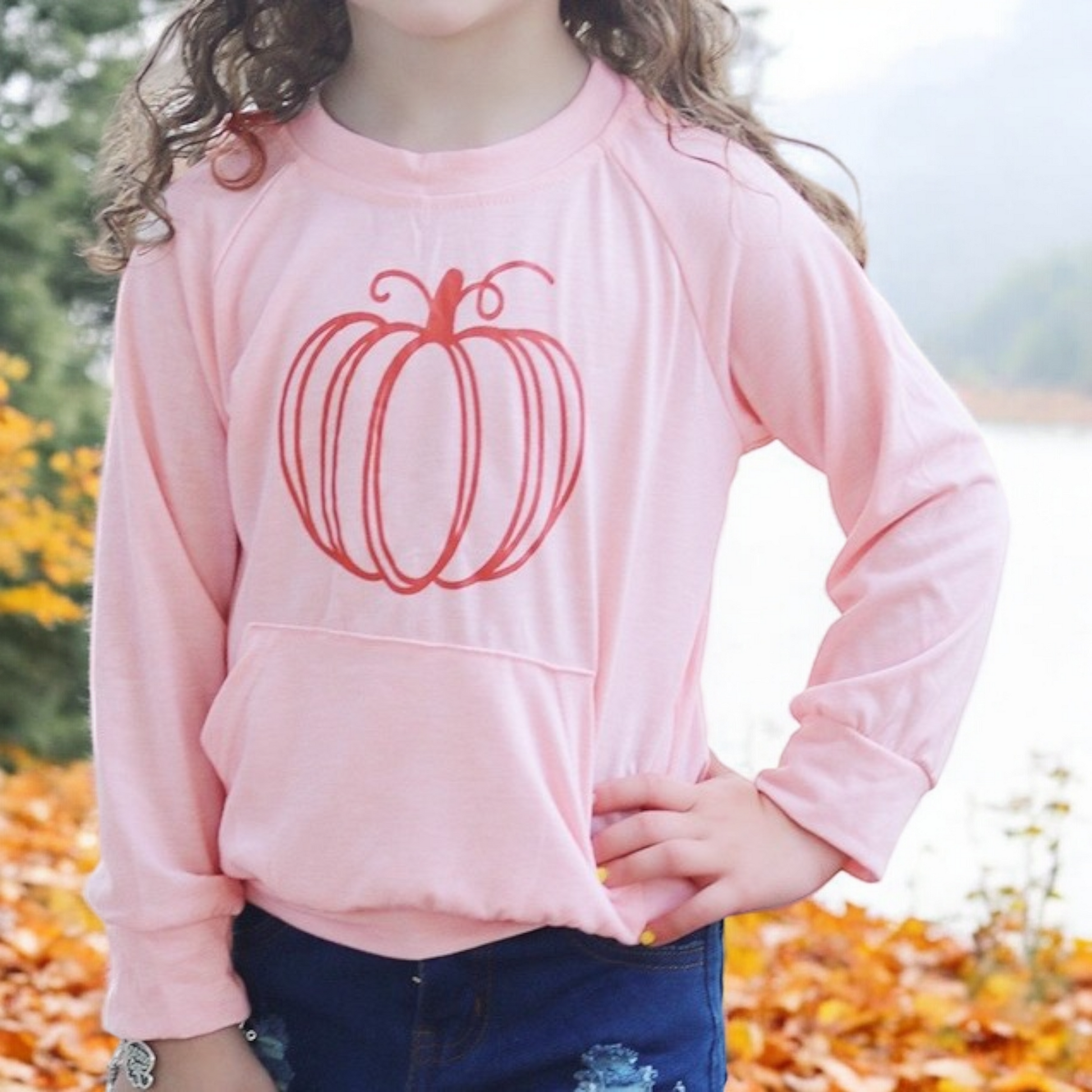 child wearing pink pumpkin sweatshirt with front pumpkin graphic outdoors in fall season