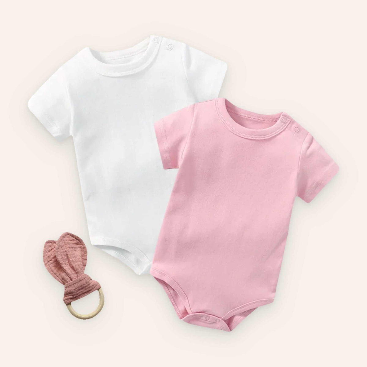 Baby bodysuit set with white and pink bodysuits and rabbit ear teether on wooden background.