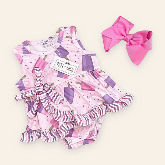 Popsicle Princess Romper with pink and purple popsicle print, ruffle details, and a side bow, shown on a light wood background.